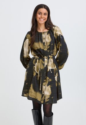 Femme souriante portant une robe à boutons avec motif abstrait noir et or, ceinturée à la taille, des collants noirs et des bottes noires montantes jusqu'aux genoux.