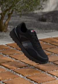 Black athletic shoe featuring mesh and synthetic materials, textured surface, a logo on the tongue, and reflective accents on the side.