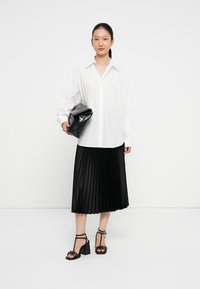 Hvid oversized knap-op skjorte, sort plisseret midi-nederdel, sorte hælede sandaler med remme og en sort clutch. Ensfarvet baggrund.