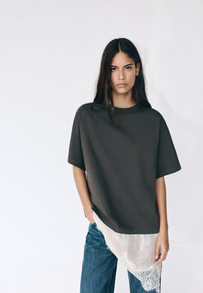 Junge Frau mit langen dunklen Haaren, die ein lockeres dunkelbraunes Oberteil mit weißen Spitzenakzenten und blaue Jeans trägt, stehend vor einem schlichten weißen Hintergrund.