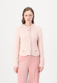 Vaaleanpunainen neulecardigan, jossa on pyöreä pääntie, nappikiinnitys ja teksturoitu pinta. Siinä on kaksi etutaskua napitus yksityiskohdilla.