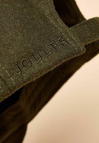 Olīvu zaļas filca izstrādājums ar šuvju akcentiem. Redzams iespiests "JOULES" logotips. Mīksta tekstūra, rādot vienkāršu un funkcionālu dizainu.