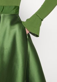 Mano en el bolsillo de una brillante falda de satén verde, combinada con un suéter verde acanalado a juego que presenta puños acampanados y una costura decorativa.