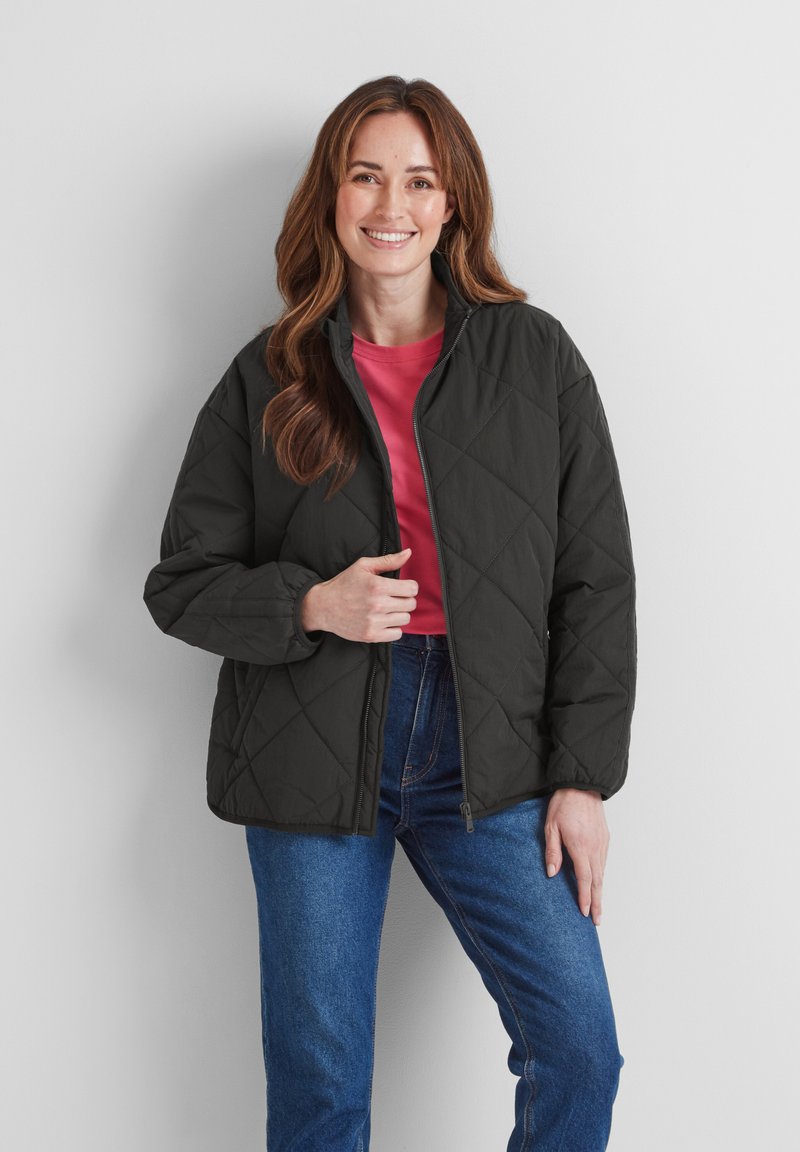 Black quilted jacket with a standing collar, zip closure, and side pockets, worn over a pink shirt and paired with blue jeans.