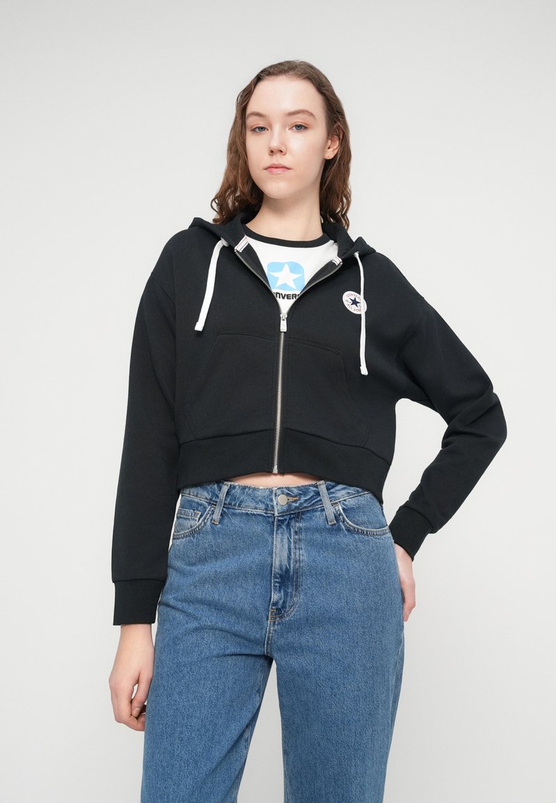 Sort cropped hættetrøje med lynlås foran, hvide snore og et logo-patch. Båret over en blå grafisk t-shirt og kombineret med blå jeans.