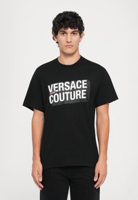 Sort bomulls-T-skjorte med korte ermer, med fet hvit og rosa tekst: "VERSACE JEANS COUTURE" over brystet.