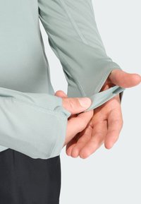 Hænder, der justerer manchetterne på en lyseblå skjorte med lange ærmer og tommelfingerhuller, og viser det teksturerede stof og syningsdetaljer.