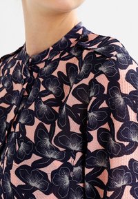 Kortärmad blus i rosa och marinblå med blomsterdesign, texturerat tyg, knappdetaljering och klassisk krage.