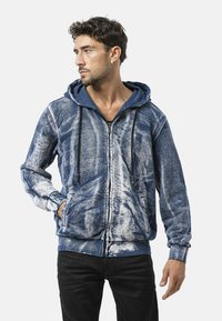 Blauwe hoodie met ritssluiting en abstracte witte patronen, voorzien van een capuchon, voorzakken en donkere trekkoorden. Gemaakt van zacht stof.