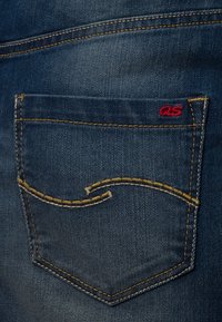 Tylnia kieszeń dżinsów z niebieską tkaniną, żółtym przeszyciem i czerwonym logo "QS". Charakteryzuje się wyraźnym, zakrzywionym szwem.