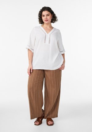 Kobieta ubrana w białą bluzkę z dekoltem w serek i koronkowymi detalami, brązowe, szerokie spodnie w paski oraz brązowe sandały, stojąca na tle białej, jednolitej ściany.