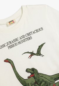 Camiseta blanca de algodón con gráficos de dinosaurios verdes, incluyendo un T-rex y un pterosaurio, con texto negro que dice "Monstruos Clásicos, Jurásicos y del Período Cretácico."