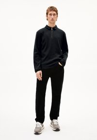 Schwarzes Langarm-Polo mit Reißverschlusskragen, aus strukturiertem Stoff, kombiniert mit schwarzen Cordhosen und grauen Sneakers.