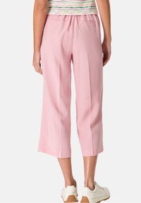 Žena v krátkých růžových kalhotách, pruhovaném tričku a bílých teniskách, zobrazená zezadu na jednobarevném pozadí.