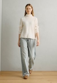 Lang kremfarget ribbestrikket genser med høy krage, båret over lyseblå vide jeans med dekorativ søm på siden, kombinert med brune sko.