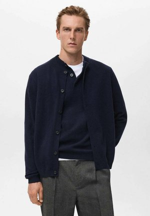 Cárdigan navy de botones hecho de lana con cuello redondo, tejido texturizado y puños acanalados, superpuesto a una camisa blanca.