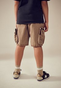 Beežid cargo-tüüpi lühikesed püksid küljekottidega, seljas on mustad New Balance tossud ja beežid sokid, millel on valged triibud. Näha on must T-särk.