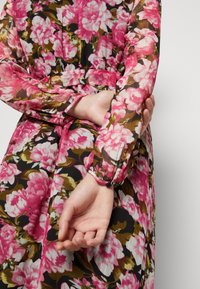 Blommig klänning med långa, genomskinliga ärmar i rosa och vita blommor på en svart bakgrund. Har en åtsittande midja och knäppta manschetter.