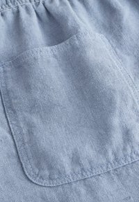 Poche en tissu bleu clair avec couture visible sur matériau en lin texturé, ceinture élastique froncée au-dessus de la poche.