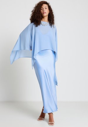 Donna con capelli ricci che indossa un abito azzurro fluente con maniche larghe e una collana vistosa, in piedi su uno sfondo bianco.