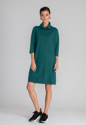 Tefarbene, knielange Kleid mit Trichterkragen und Dreiviertelärmeln, kombiniert mit schwarzen Sportschuhen. Glattes, mittelschweres Material.