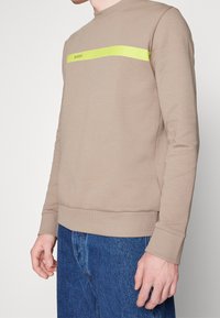 Beige sweatshirt i mjuk bomull, med ett klart gult horisontellt rand och logotyp. Ribbstickade muddar och nederkant, i kombination med blå denim.