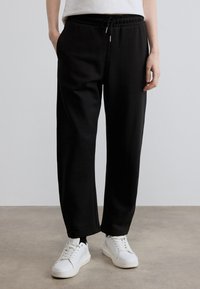 Μαύρο παντελόνι jogging από μαλακό υλικό με ελαστική μέση και κορδόνι. Φορεμένο με λευκά αθλητικά παπούτσια, με χαλαρή εφαρμογή.