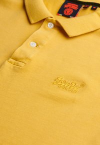 Żółta koszulka polo wykonana z teksturowanej tkaniny z trzema guzikami przy zapięciu oraz haftowanym logo "SuperDry" na lewym kawałku klatki piersiowej.