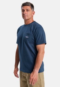 Navyblaue Baumwoll-T-Shirt mit rundem Halsausschnitt, kurzen Ärmeln und einer Brusttasche auf der linken Seite, die ein kleines Logo zeigt. Einfaches Design und Textur.