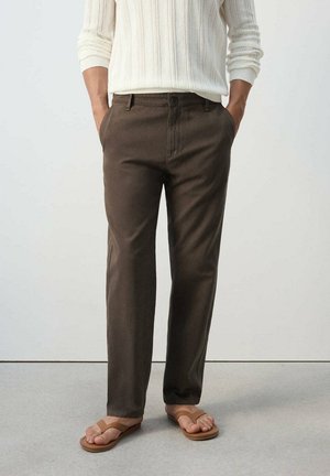 Hombre con pantalones marrones casuales, un suéter de punto blanco de manga larga y sandalias marrones, de pie con las manos en los bolsillos.