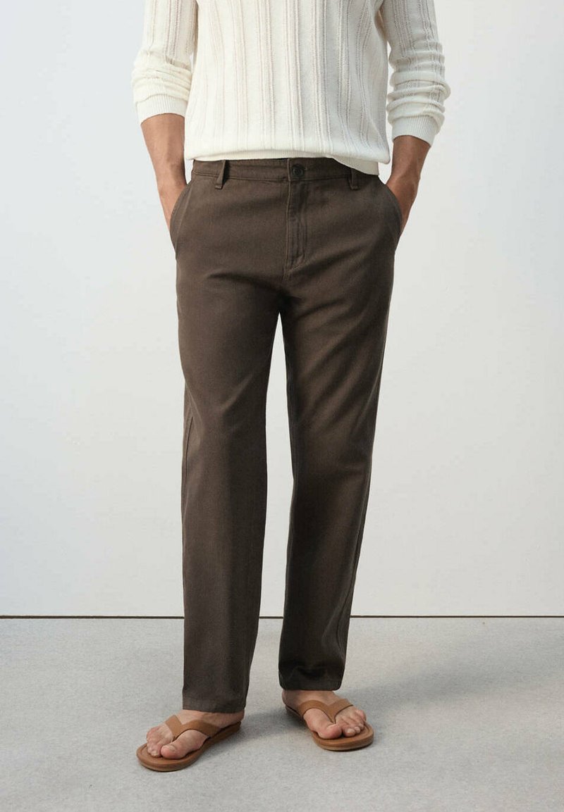 Homme portant un pantalon marron décontracté, un pull en maille blanc à manches longues et des sandales beige, debout les mains dans les poches.