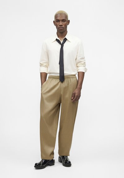 Άνδρας με κοντά ξανθά μαλλιά, που φορά λευκό πουκάμισο, μαύρη γραβάτα, φαρδιά καφέ παντελόνια και μαύρα γυαλισμένα παπούτσια, στέκεται μπροστά σε απλό λευκό φόντο.