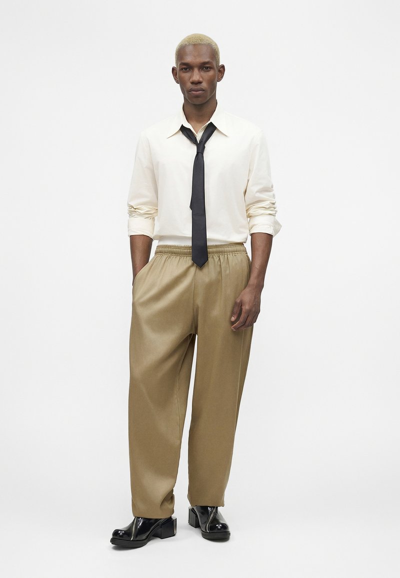 Homme aux cheveux blonds courts portant une chemise blanche, une cravate noire, un pantalon beige ample et des chaussures noires vernis, debout devant un fond blanc uni.