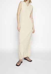 Robe maxi sans manches, décolletée en V, de couleur crème, en tissu tricoté, avec une coupe légèrement cintrée et un ourlet côtelé. Associée à des sandales noires à lanières.