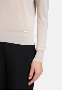 Pull léger beige avec une texture douce et transparente, des poignets côtelés et un subtil accent de logo près de l'ourlet, associé à un pantalon noir.