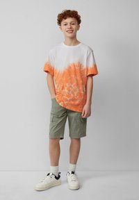 Korte mouwen T-shirt in een witte en oranje tie-dye patroon, gecombineerd met olijfgroene cargoshorts en witte sneakers met zwarte accenten.