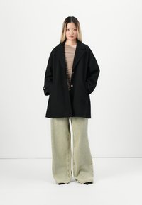 Black oversized coat with two buttons and side pockets, paired with beige fitted top and wide-leg light green jeans. Smooth texture.