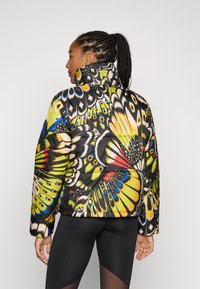 Casaco acolchoado com um estampado colorido de borboletas em amarelo, verde e azul. Gola alta e base preta. Modelo a usar leggings pretas.