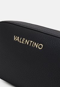 Černá kožená taška s texturou a zakulaceným tvarem. Obsahuje zlaté logo "VALENTINO". Hladký zipový uzávěr na vrchu.