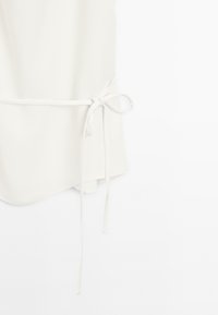 Hvid indpakningstop lavet af glat stof med et båndlukning, der har en buet kant og delikate taljebånd for en sikker pasform.