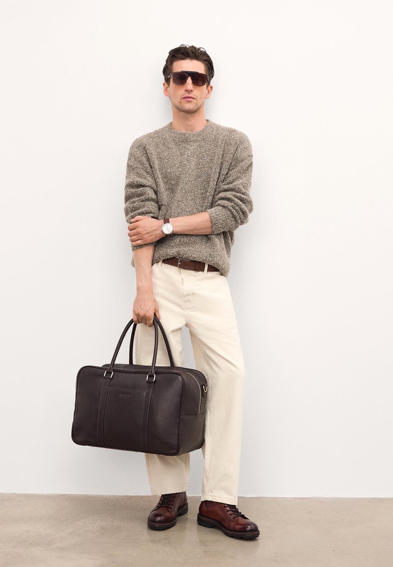 Braunā ādas soma ar divām rokturiem, apvienota ar teksturētu bēšu džemperi, krēmkrāsas biksēm un brūnām šņorējamām zālēm. Gaismas fons.