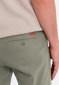 Pantalon vert avec une poche arrière dotée d'un bouton en métal et d'une étiquette en cuir marron. Le tissu a une texture lisse et une coupe ajustée.