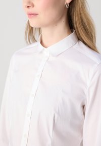 Bílá košile s knoflíky a špičatým límcem, která se pyšní jemným vyšívaným logem na prsou a hladkou, lehkou teksturou látky.