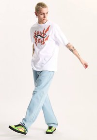 Weißes T-Shirt mit orangefarbenem Grafikdesign, hellblaue Jeans und schwarze Sneaker mit neongrünen und pinken Akzenten.