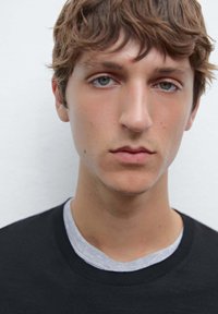 Black sweatshirt with a gray crew neck collar, worn by a person with short, wavy brown hair and light blue-green eyes, against a neutral background.