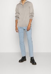 Maglione beige lavorato a maglia con collo a zip parziale, abbinato a jeans a gamba dritta azzurri chiaro e stivaletti neri alla caviglia.