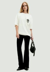 Valkoinen ylisuurikokoinen t-paita, jossa on graafinen kuvio rinnassa, yhdistettynä mustiin levennettyihin housuihin. Malli pitää pientä mustaa laukkua ja käyttää mustia ballerinoja.