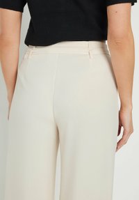Pantalones ajustados color beige claro con una textura suave, detalles de costura en la cintura y bolsillos laterales discretos. Vista posterior, diseño minimalista.