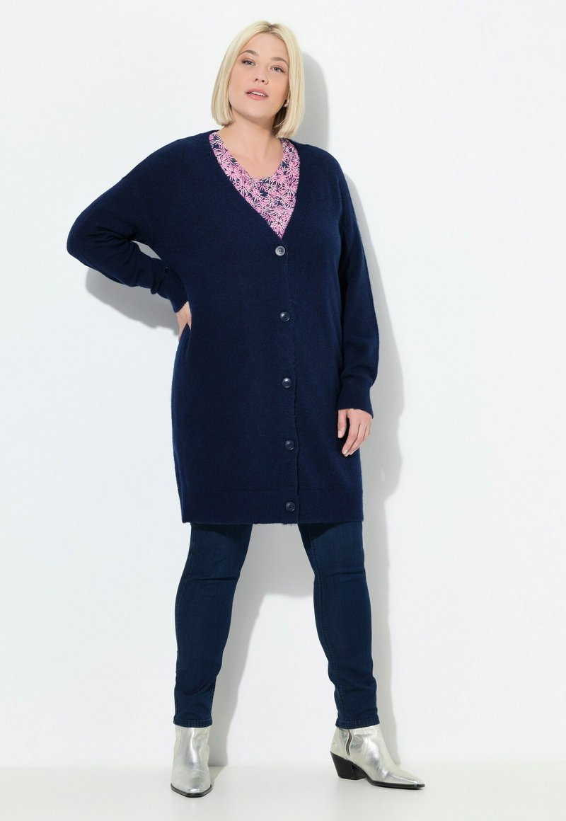 Marinblå lång cardigan i mjukt tyg, med v-ringning, knäppning och två framfickor, tillsammans med mönstrad topp och jeans.