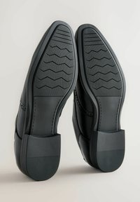 Pantofi formali negri cu tălpi din cauciuc texturat și modelat și toc jos, arătați din partea inferioară pe un fundal neutru.